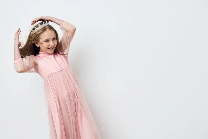 Cheerful girl in a pink dress wearing a crown, showcasing joy and playfulness against a soft light background, perfect for children s fashion or celebration concepts