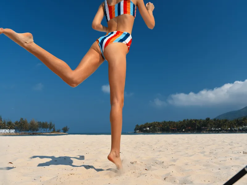 A person in striped bikini runs along a sunlit sandy beach under a clear blue sky, capturing dynamic movement and vibrant summertime energy.