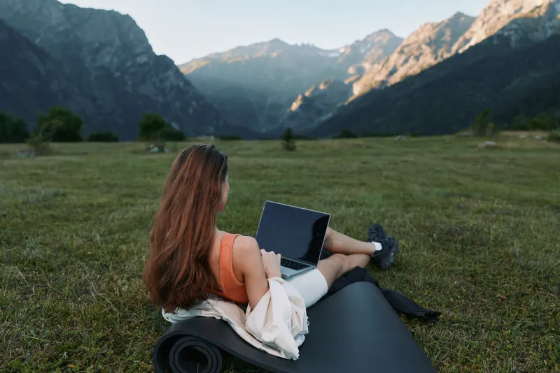 A person sits on a yoga mat in a scenic valley, working on a laptop. The setting is tranquil and inspiring, highlighting remote work in nature.