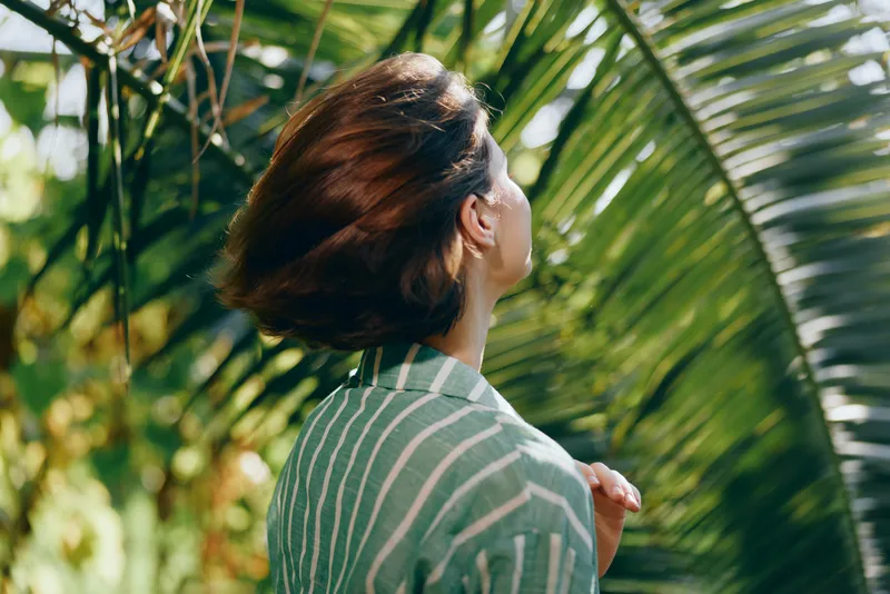 Portrait of a person seen from behind, looking up amid lush palm leaves with warm sunlight, conveying curiosity and nature-inspired calm.