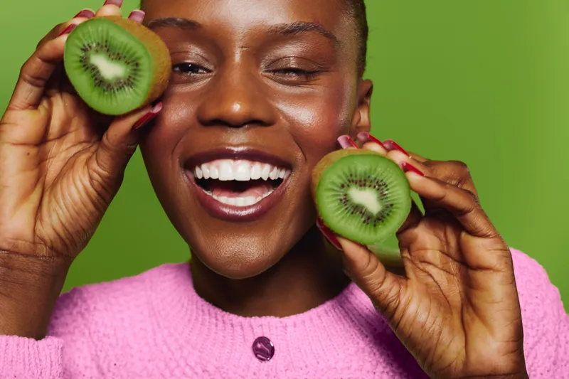 Happy African woman with dark skin holds two halves of fresh kiwi fruit near face. Close-up portrait with bright green background. Concept of healthy food, nutrition, and positive lifestyle.