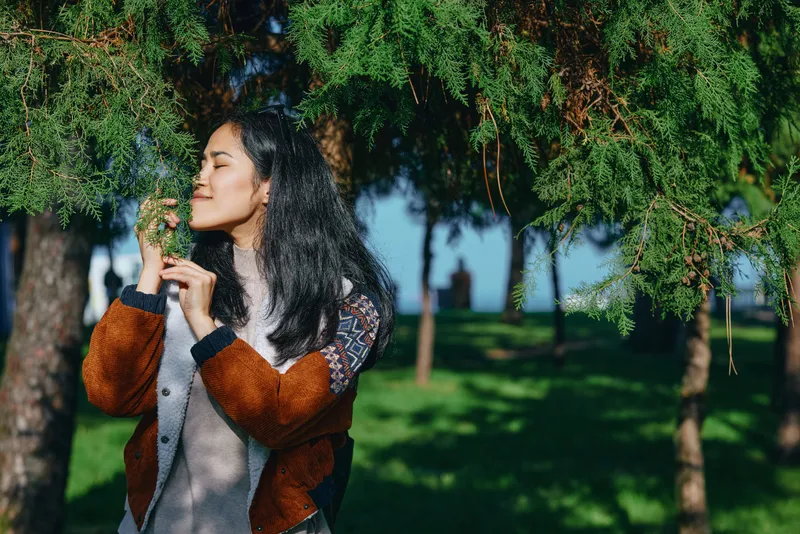 Nature, woman, outdoors, park, greenery, tree branches frame a candid moment as she smells a leaf, enjoying fresh air, tranquil scenery, and peaceful park mood