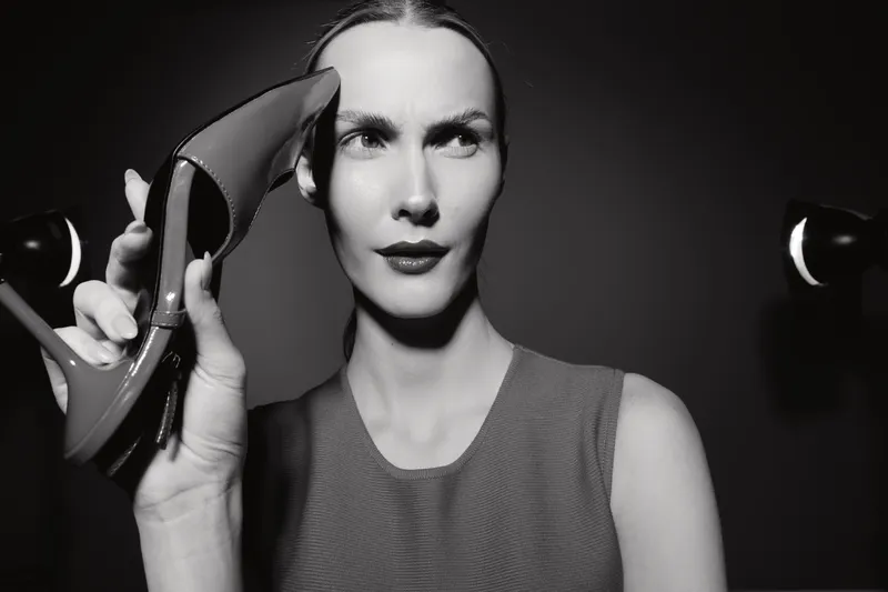 White woman with fair skin holds a high heel shoe near her face, looking serious in black and white studio lighting. Concept for fashion, style, and elegance.