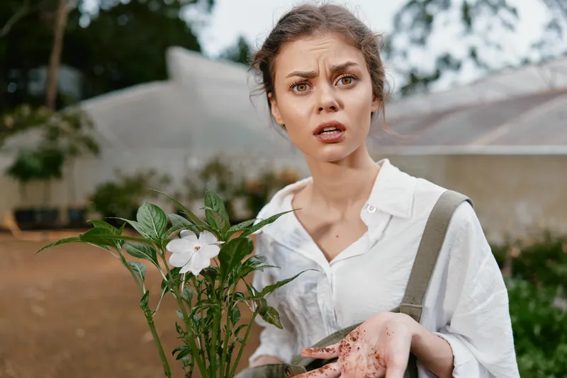 Young woman with messy hands, surprised expression, holding a plant in a garden, showcasing uncertainty in gardening, perfect for eco friendly and gardening themes