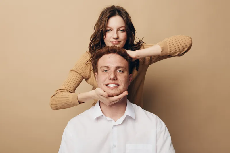 Smiling young man in white shirt with woman behind him framing his face with hands on beige background. Concept of friendship, happiness, casual style, and positive emotions in studio.