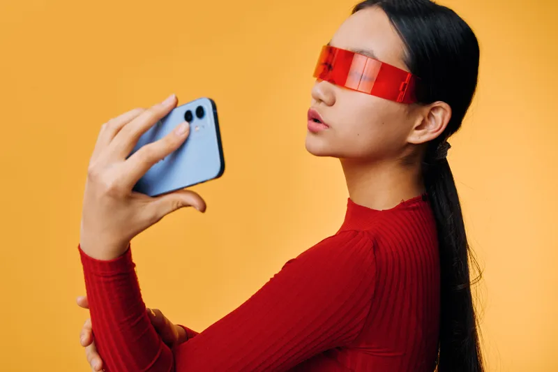 Woman in red sweater and futuristic red glasses taking selfie with smartphone on orange background, modern technology and stylish portrait for social media content creation.