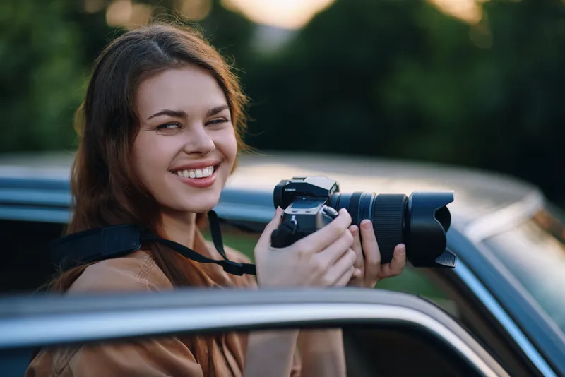Happy young woman photographer capturing moments with a camera, smiling while sitting in a car; natural background in soft colors, showcasing joy and creativity.