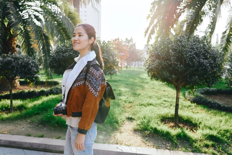 Woman with backpack strolling through a sunlit park, casual fashion and walking in greenery under bright day light, an outdoor lifestyle shot for travel and nature themes