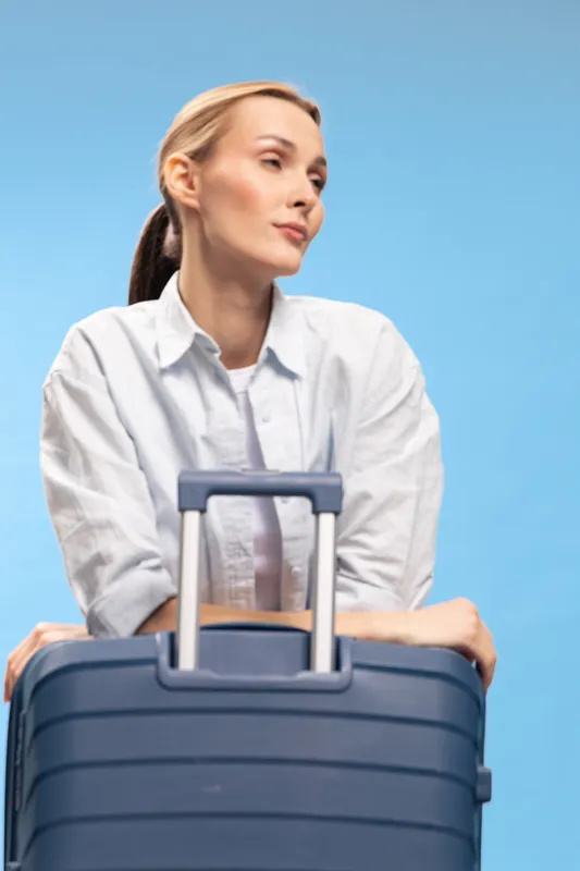 Young woman in casual white shirt leans on blue suitcase handle, looking away thoughtfully on blue background. Concept of travel, waiting, and planning a trip with luggage.