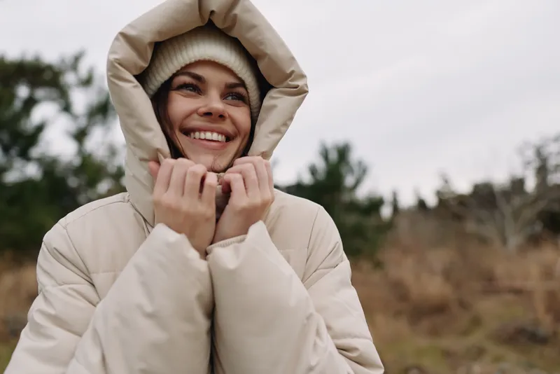 Winter scene with a smiling woman wearing a thick coat and hood, outdoors in cool weather, conveying warmth and comfort as she gazes with delight.