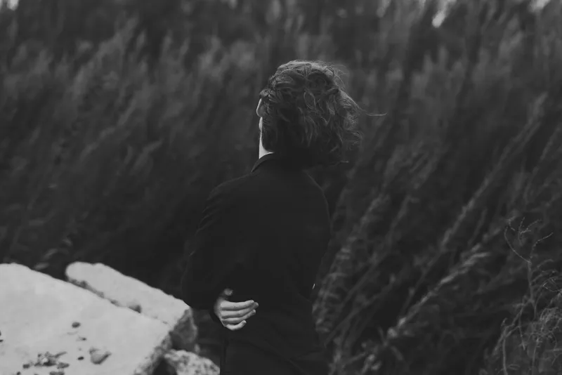 Silhouette of a woman with her back turned, standing near a rocky shore and tall grasses, evoking solitude, reflection, and quiet nature in monochrome.