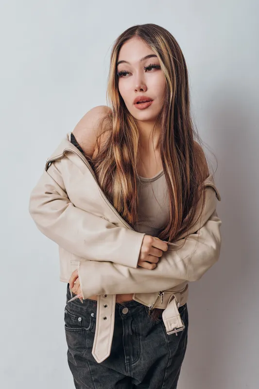 Young woman with long hair wearing light jacket and tank top posing against white background. Casual fashion style, portrait with thoughtful expression and natural makeup in studio.
