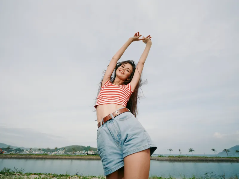A carefree young woman stands outdoors with arms raised, wearing a striped crop top and denim shorts, capturing a bright summer mood against a blue sky.