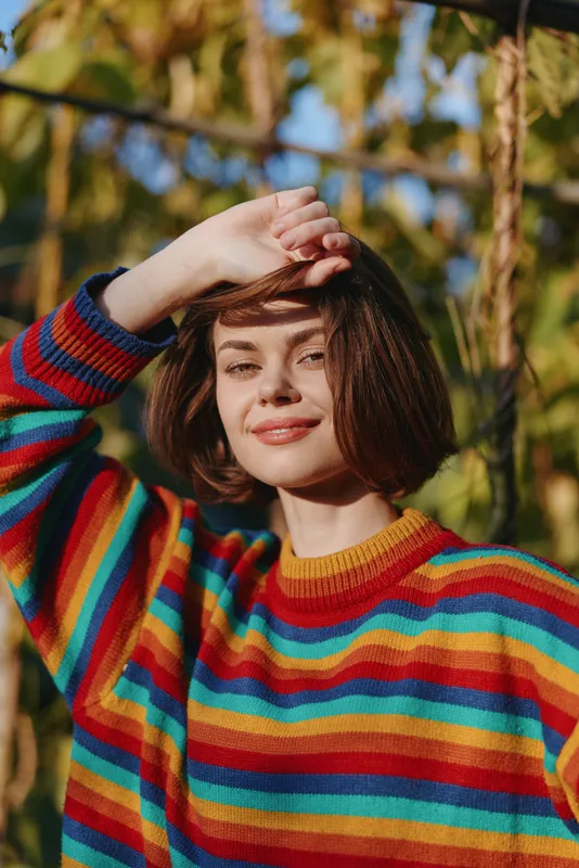 Young woman in her early 20s wearing a colorful striped sweater, smiling portrait outdoor in golden autumn light, hand shielding eyes, casual fashion lifestyle headshot.
