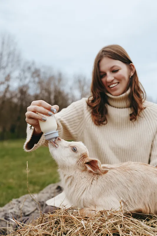 Young woman in a beige sweater happily feeds a baby goat with a bottle outdoors in a natural rural setting during a calm day.