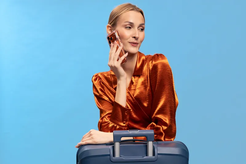 Young woman in shiny orange blouse holds phone to ear while resting arms on suitcase handle. Blue background highlights travel concept, communication, and stylish business trip preparation.