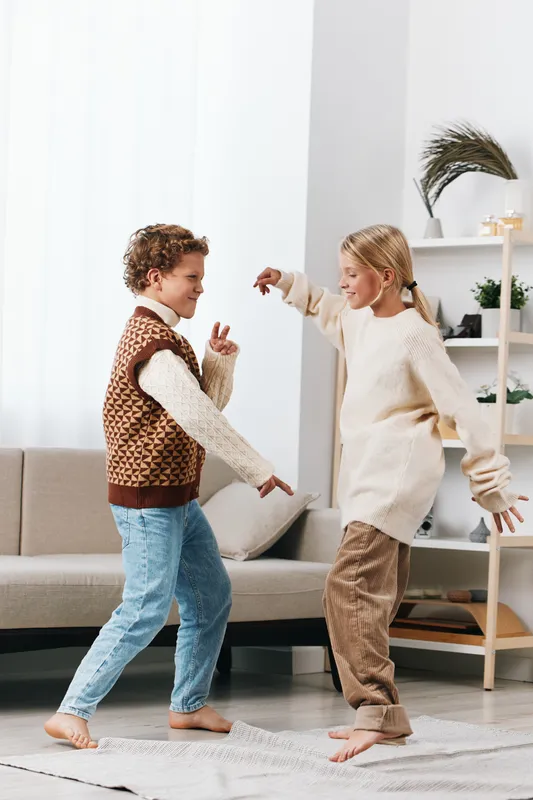 Two children dance in a bright living room, barefoot and smiling, captured in a candid moment of playful movement and carefree energy inside a cozy home.
