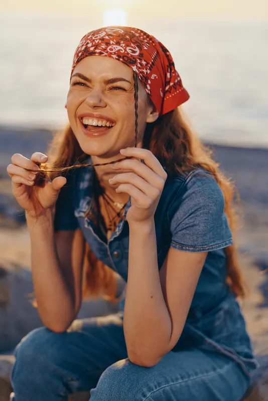 Lifestyle woman in modern boho-western denim street style wearing a red bandana and smiling warmly with a film color effect on a sunny beach background.