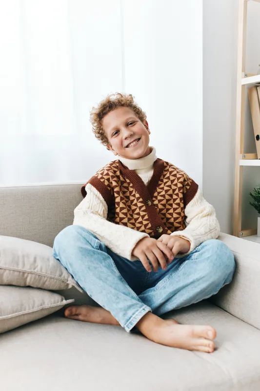 Smiling young boy sits cross legged on a light sofa, wearing a brown patterned knit vest over a cream sweater and jeans, in a bright cozy living room.