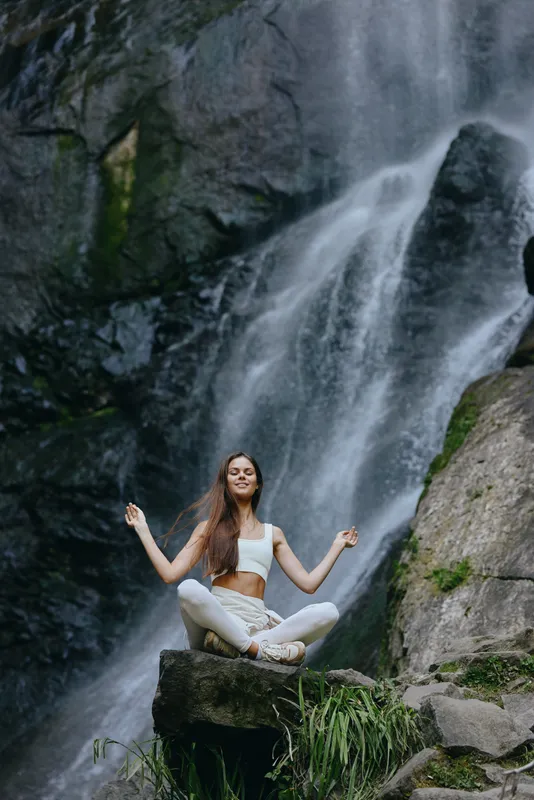 young woman meditating near waterfall in nature, peaceful, calm, yoga practice by rocks and flowing water with green plants
