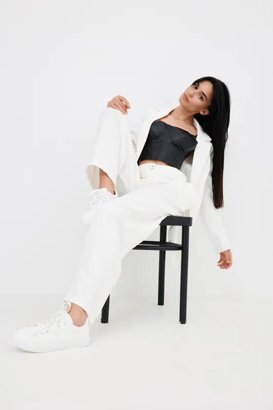 young woman posing on black chair wearing white trousers, white jacket, black crop top, and white sneakers against plain white background, stylish and minimal look