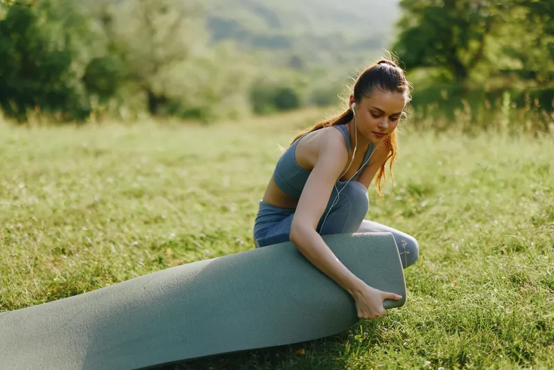 Young woman practicing yoga outdoors, rolling up a mat in green grass, showcasing a healthy lifestyle, fitness, and tranquility in nature
