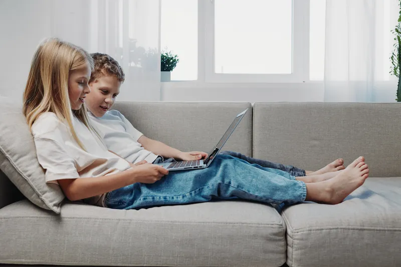 Two children sitting on a grey sofa using a laptop together in a bright living room. Concept of learning, technology, childhood, education, and family bonding at home.