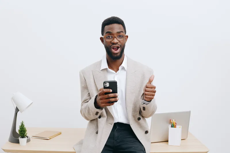 Confident young African American man wearing glasses and beige blazer using smartphone gesture thumbs up, standing in modern office with minimalist decor white background corporate communication conce