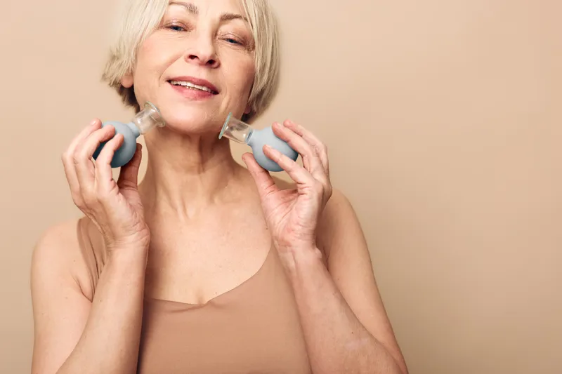 Mature woman with short gray hair applies facial massage cups on cheeks in beige tank top. Skincare concept for anti aging, self care, wellness, and natural beauty with soft neutral background.
