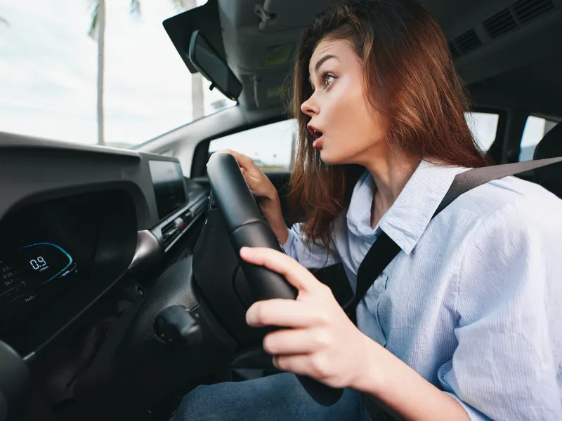 Женщина ведет машину, держа одну руку на руле, и удивленно открывает рот, глядя на что-то на приборной панели