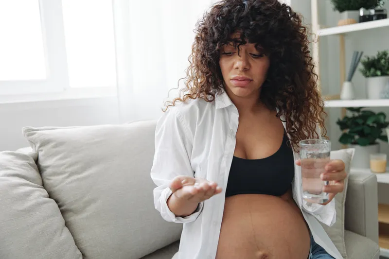 Беременная женщина улыбается и чувствует счастье, принимая таблетки и витамины для беременных: в одной руке у неё таблетки, в другой — стакан воды, она сидит дома на диване