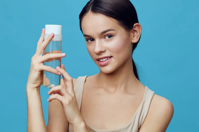 Young woman holding a skincare bottle, smiling with natural makeup, on a bright blue background, perfect for wellness and beauty concepts