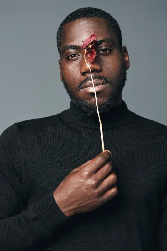 African man with dark skin holding dried flower in front of face in creative conceptual visual style. Portrait of male model wearing black turtleneck on gray background.