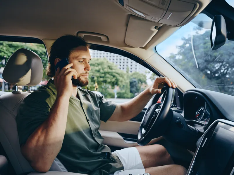 A man sits behind the wheel, driving while talking on a mobile phone. He wears casual clothes, sunlight spills inside the cabin, focused on the road ahead.