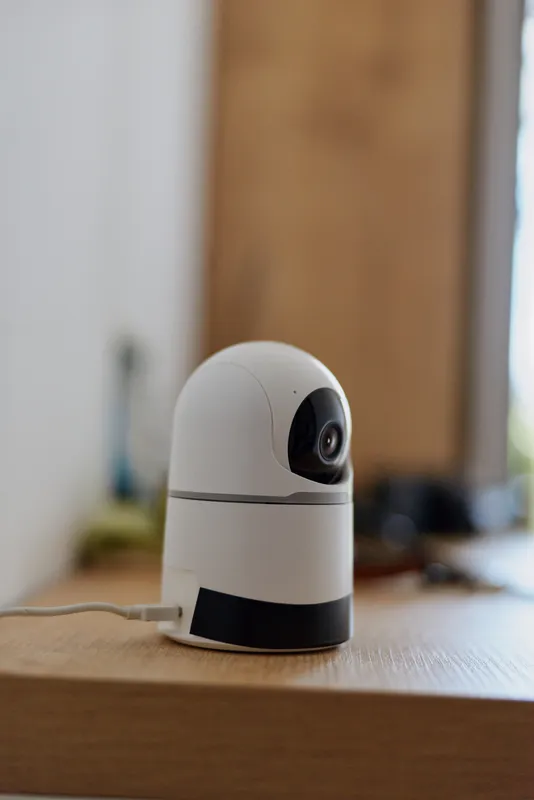 Close up of a small white indoor webcam on a wooden table, modern security device in a minimal home setting, soft natural light and calm atmosphere, everyday tech