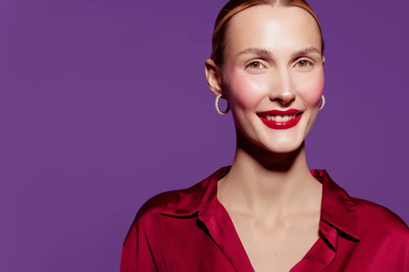 Smiling woman with red lipstick and hoop earrings wears red blouse against purple background. Portrait of confident female model with slicked hair and natural skin glow.