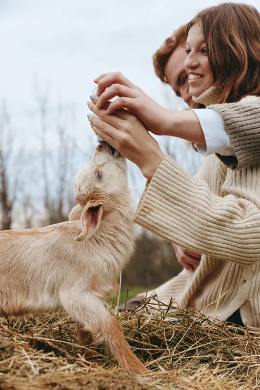 Smiling woman with a curious goat kid in an open field, outdoor lifestyle moment. Natural light highlights gentle care, playful interaction, and a warm connection to nature. In quiet.