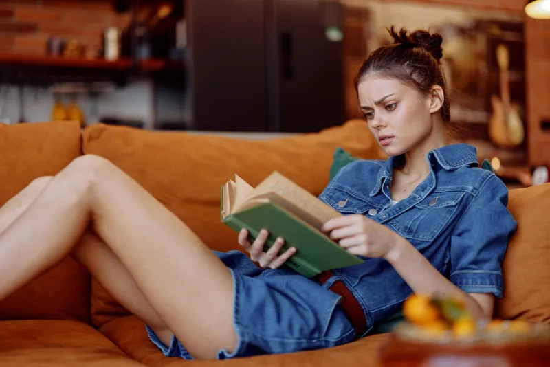 Молодая женщина в джинсовом платье читает книгу на диване в туфлях на высоком каблуке, приятно проводя время дома