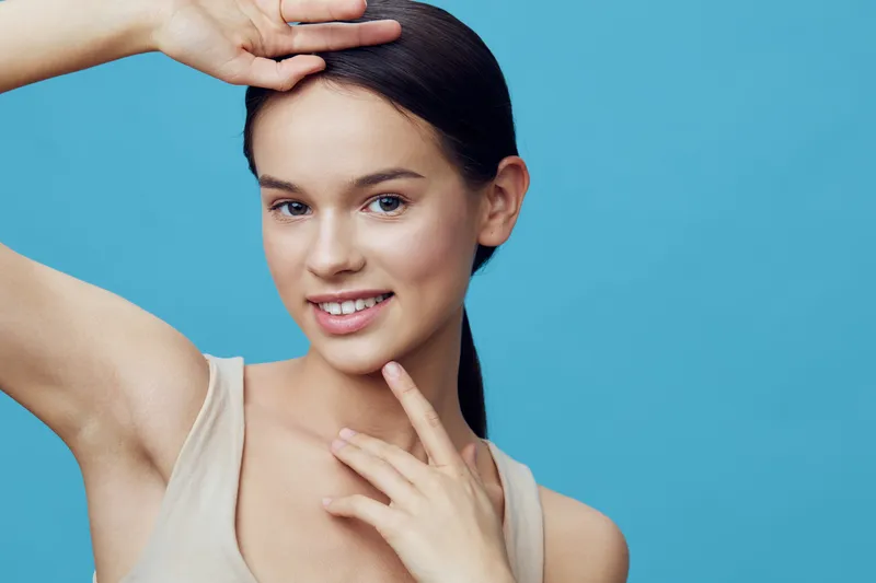 Young model with clear skin touching her face, showcasing a joyful expression against a vibrant blue background, emphasizing beauty and self care