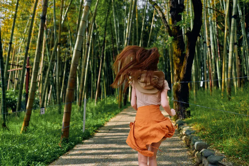 Woman running through a bamboo path in an orange skirt, back view capturing motion and flowing hair. Forest trail with sunlight and green nature evokes freedom and travel.