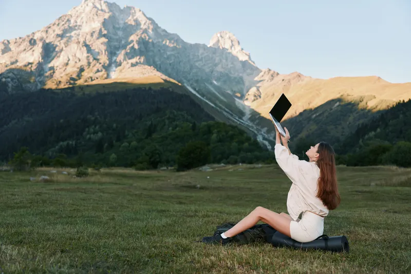 A joyful graduate sits on the grass in a mountain meadow, cap raised high as a diploma catches sunlight, exuding achievement, pride, and a sense of freedom.