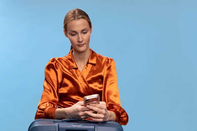 Young woman in orange satin blouse sitting with suitcase looks at smartphone on blue background. Concept of travel preparation, communication, and modern lifestyle.