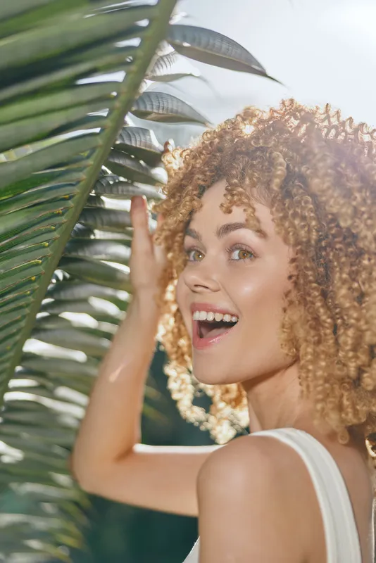 Smiling woman with curly hair enjoys tropical outdoor atmosphere surrounded by green palm leaves and bright natural sunlight, radiating joy and warmth.