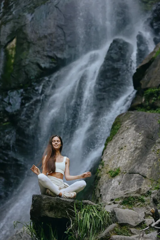 Woman meditating near waterfall in white athletic wear, practicing yoga outdoors on rock surrounded by nature and flowing water, peaceful and serene atmosphere.