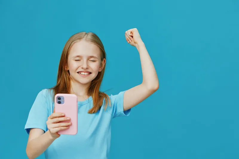 Happy girl celebrating success with mobile phone against a blue background, wearing a light blue t shirt, showcasing joy and excitement