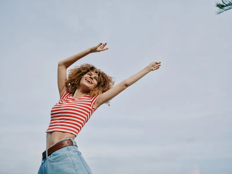 A joyful young woman dances outdoors with arms raised under a clear blue sky, wearing a striped red and white crop top and light denim jeans.