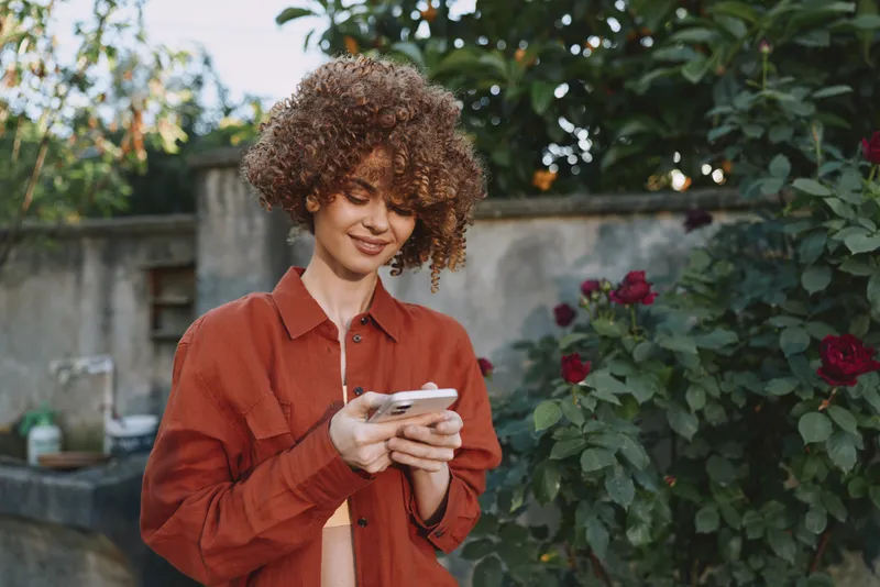 A cheerful woman with curly hair in a red shirt stands in a sunlit garden, eyes on a smartphone. Casual lifestyle scene, outdoor setting, everyday moment, candid fashion