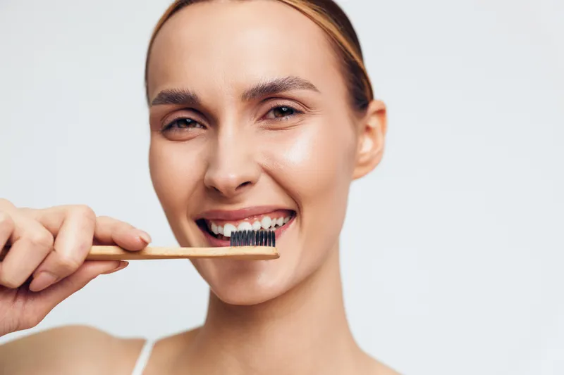 Young woman brushing teeth with bamboo toothbrush, smiling gently, clear skin, natural light, dental hygiene routine, clean white background, close-up portrait
