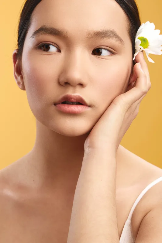 asian woman with a delicate flower behind the ear, gazing softly to the side in a studio portrait, warm background, showcasing natural beauty and serene elegance.