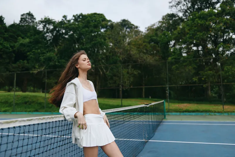 Young athletic woman in a sporty outfit, enjoying a moment of tranquility on a tennis court surrounded by lush greenery and vibrant colors, showcasing the joy of an active lifestyle